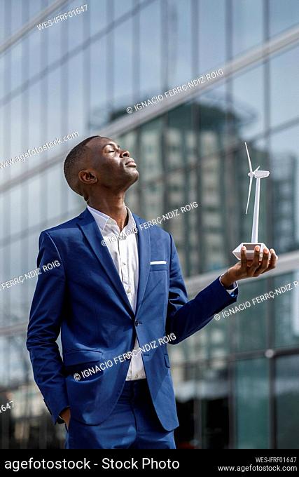 Engineer with eyes closed holding wind turbine model in front of modern building