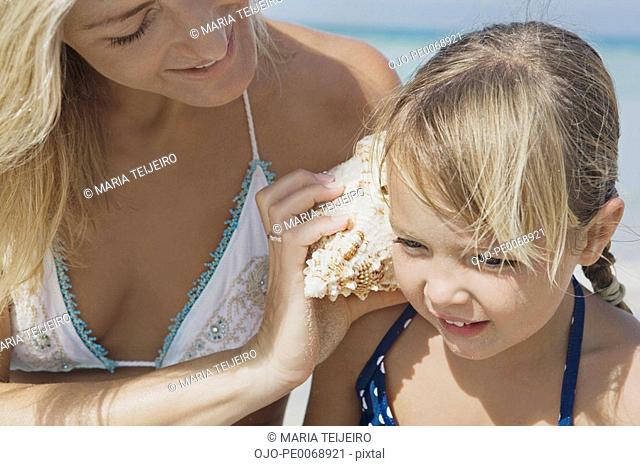 Mother holding shell up to daughter’s ear on beach