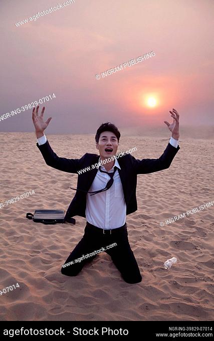 Frustrated businessman with arms outstretched kneeling in the desert at sunset