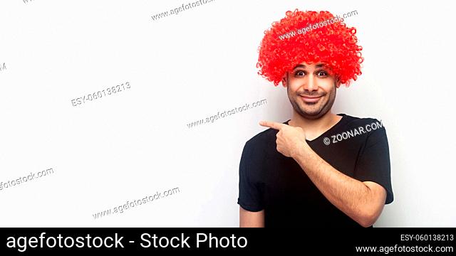 Portrait of stylish funny happy man with red wig smiling and pointing to the side, showing empty copy space for advertisement, freespace