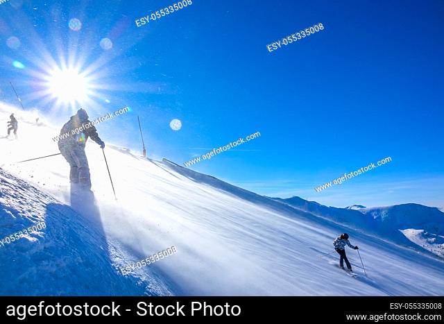 Sunny weather at the ski resort. A strong wind blows snow down a steep slope. Multiple skiers