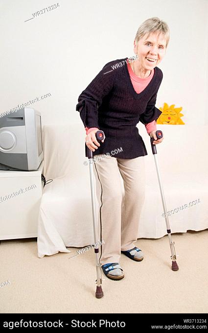 Ganzkörperansicht einer älteren Frau an Krücken im heimischen Wohnzimmer stehend nach einer Hüftoperation. Full body view of an elderly woman on crutches in...