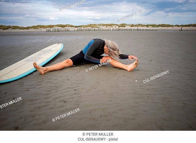 Senior woman sitting on sand, stretching, surfboard beside her