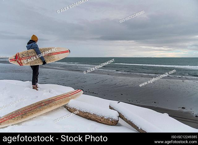 Rear view of a woman wearing a wetsuit and holding a surfboard standing on a snowy beach and looking out to sea