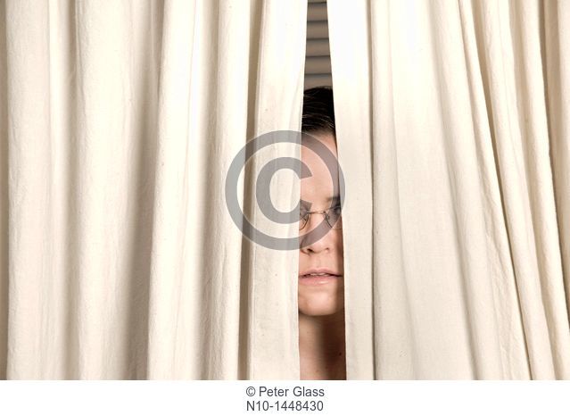 Young woman, wearing glasses, peeking out from behind two curtains