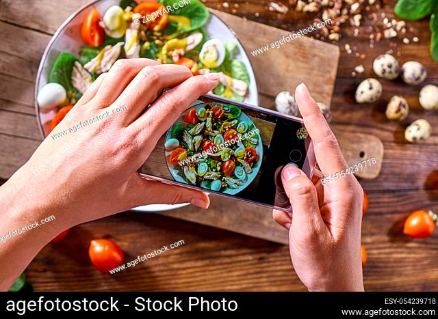 On the kitchen table on a wooden board a plate with homemade salad. Woman's hands with a phone make photo of salad