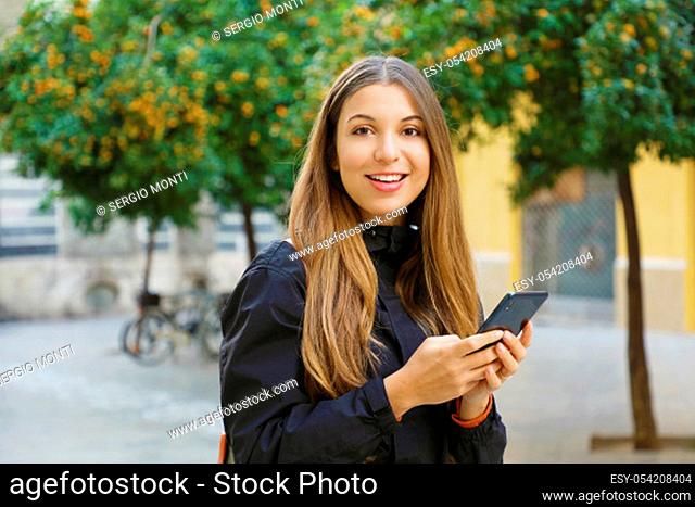 Portrait of a beautiful smiling woman using a mobile phone looking at camera in city street