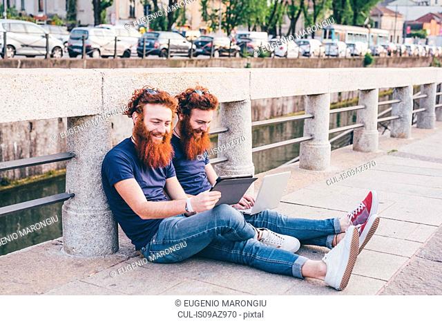 Young male hipster twins with red hair and beards browsing digital tablet on bridge
