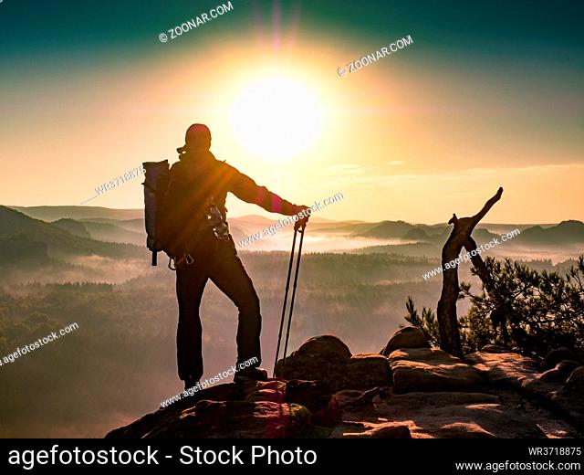 Hiker with backpack leaning on trekking poles staring into misty sunrise of Saechsische Schweiz park. Beautiful morning valley full of thick fog in orange...