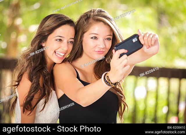 Two attractive mixed-race girlfriends using their smart cell phones for selfie outdoors