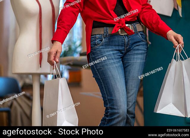 Wellbeing. Slender woman in red shirt and jeans holding shopping bags standing in sewing workshop no face