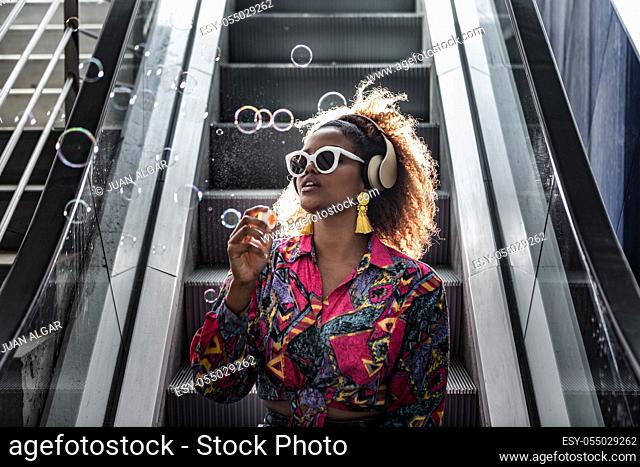 African American lady in sunglasses and colorful blouse sitting on moving staircase with open mouth while blowing bubbles in back lit