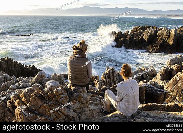 Woman and teenage girl sitting on rocky shore, looking out to sea