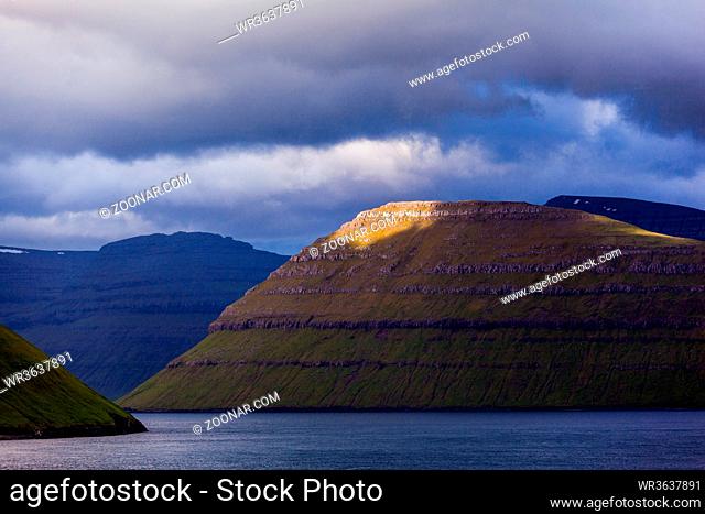 coastal line of the Faroe Islands with an overcast and dramatic sky and a patch of sun