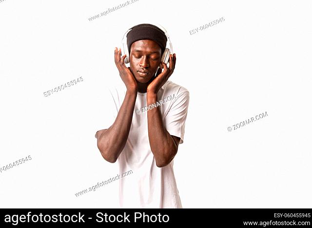 Portrait of relaxed african-american dreamy guy, enjoying listening music in headphones with closed eyes, satisfaction from sound, white background