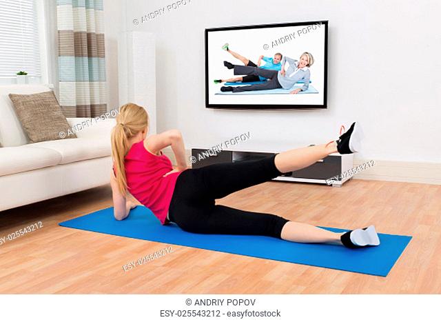Happy Woman In Sportswear With Exercise Mat Exercising In Front Of Television In Living Room