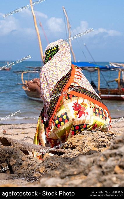 02 August 2021, Tanzania, Fumba: A woman sits in front of fishing boats on the beach. Photo: Sebastian Kahnert/dpa-Zentralbild/dpa