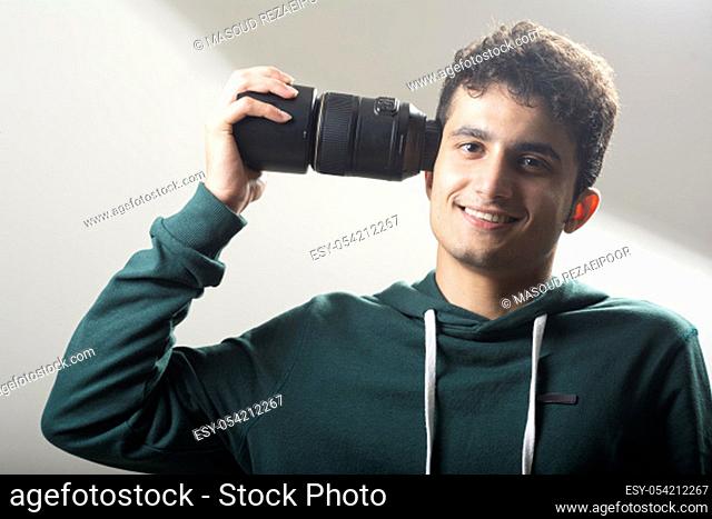 Young man wearing green pullover and holding a macro lens on the right side of his head in front of light gray background