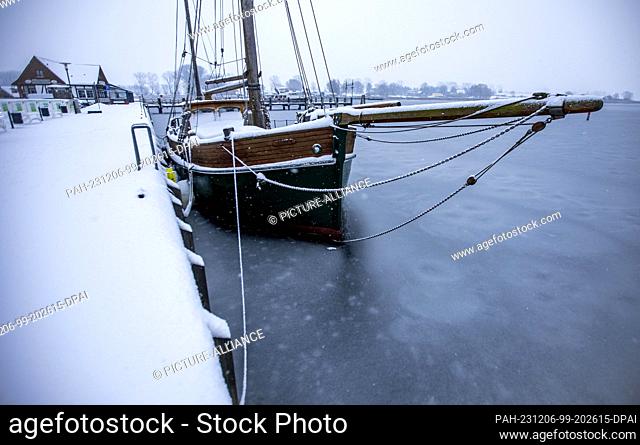 06 December 2023, Mecklenburg-Western Pomerania, Kirchdorf (poel): A historic sailing ship covered in snow is frozen in thin ice in the harbor on the Baltic Sea...