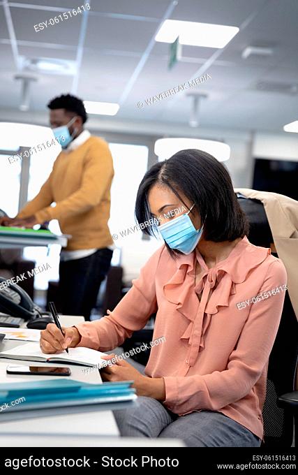 Businesswoman wearing face mask taking notes while sitting on her desk at modern office