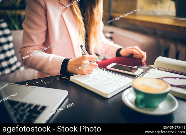 Close up caucasian woman hand on wooden table inside cafe makes notes in notebook. Subject freelancer blogger journalist at work