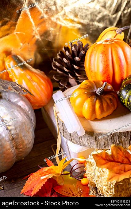 Small orange pumpkins, orange squash, pinecones, a green squash, and glue sticks for crafting on wood stacked on leaf-covered hay next to a silver pumpkin with...