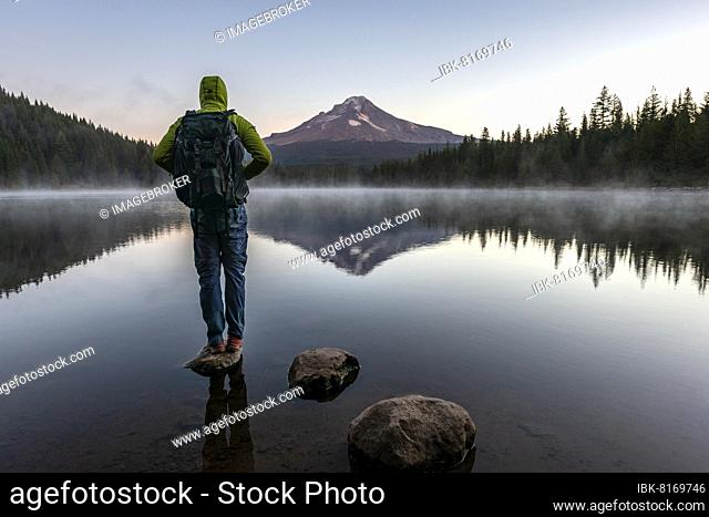 Young man standing on a stone, reflection of Mt. Hood volcano in Trillium Lake, at sunrise, Oregon, USA, North America