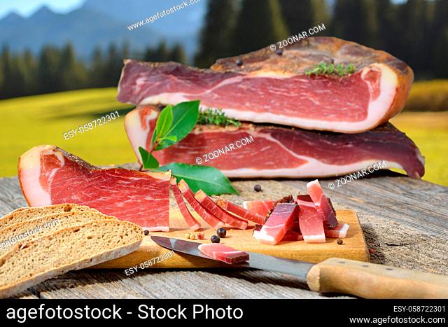 Suedtiroler Speckjause mit Vinschgerl auf einem Holztisch vor Bergen in den Alpen ? Typical South Tyrolean bacon snack with local rye bread on a rustic table in...