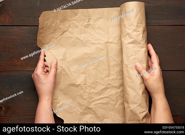 two female hands hold a rolled roll of brown craft paper on a wooden table, top view