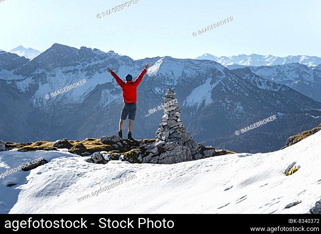 Mountaineer stretching his arms in the air, next to a cairn, in front of snow-covered mountains of the Rofan, hiking trail to Guffert with first snow, in autumn