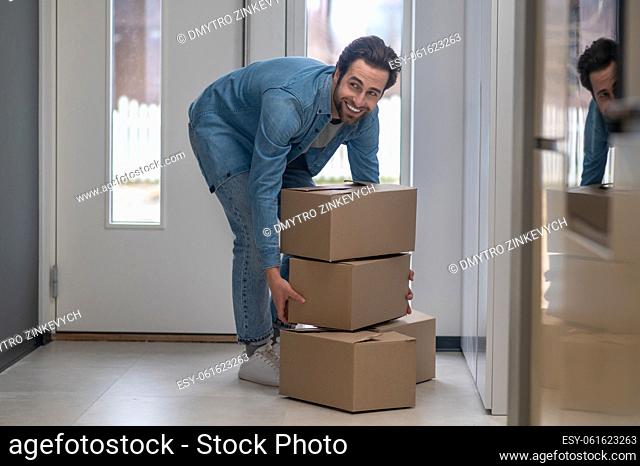 Good mood. Bearded attractive man bending over with boxes smiling to side in hallway of new apartment during day