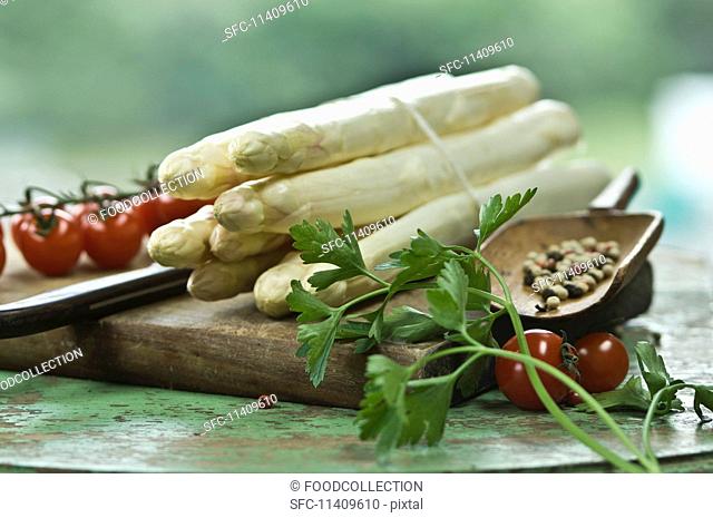 A bundle of white asparagus on a chopping board with a knife, peppercorns, parsley and cherry tomatoes