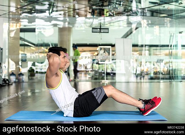 Full length side view of a fit young man sitting down on exercise mat, while doing full range crunches for the abdominal muscles at the gym
