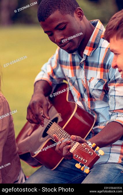 Closeup portrait of Afro-American man playing guitar while resting and relaxing with his best friends on picnic in park