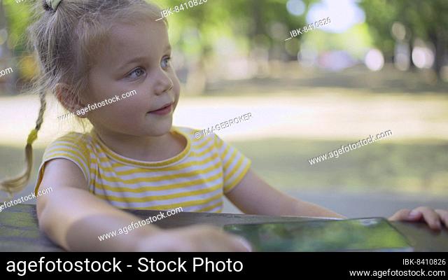 Cute little girl listens to music on a mobile phone and sings along. Close-up portrait of child girl sitting in city park and listening to music using cellphone