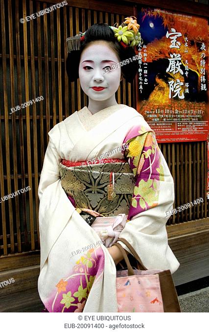 Gion District. Three-quarter standing portrait of Geisha with white facial make up and red painted lips, hair worn up with decorative pins and wearing flower...