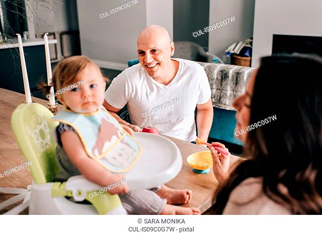 Mother and father feeding baby daughter in child seat on kitchen table
