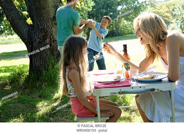 Side profile of a mid adult woman feeding food to her daughter with a boy and a mid adult man playing in the background