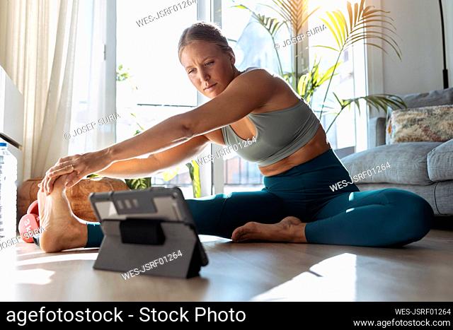 Woman exercising through digital tablet while stretching sitting on floor at home