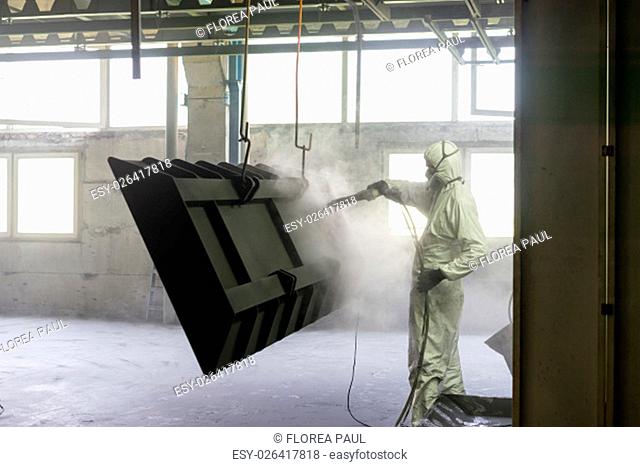 view of a worker wearing a full white protective suit and breathing mask, sand blasting a metal crate hung from a metal beam in the ceiling of an industrial...
