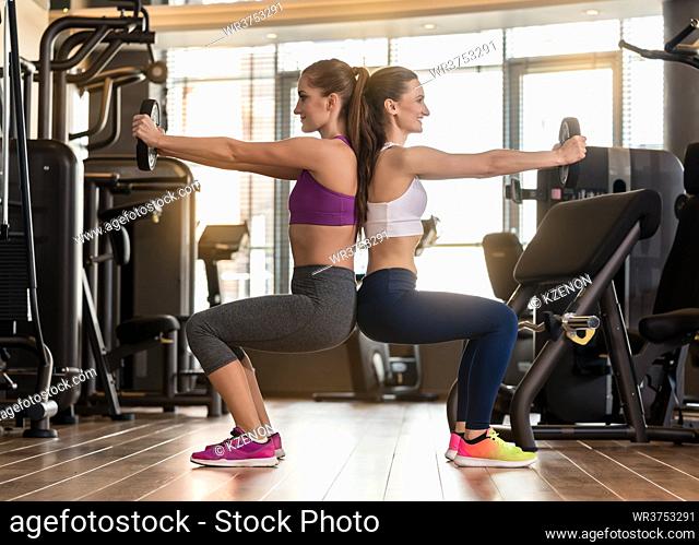 Side view of two young women smiling while exercising together back to back with weight plates in the interior of a modern fitness club