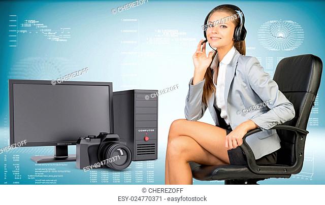 Businesswoman in headset sitting on office chair, her hand on microphone, looking at camera, smiling. Desktop computer and camera beside