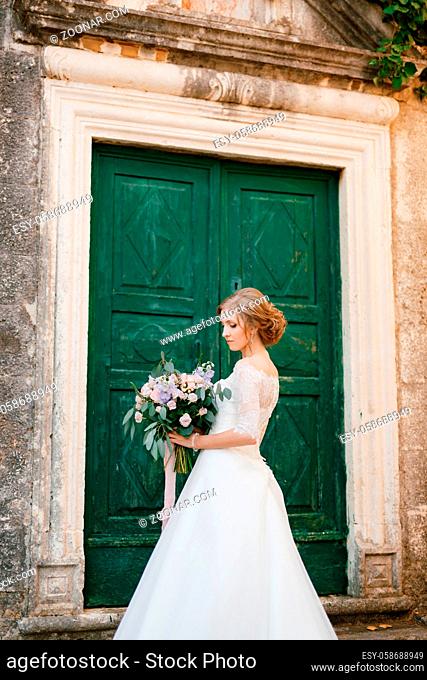 A tender bride stands with a bouquet in her hands against the background of a wooden green door of an ancient building . High quality photo