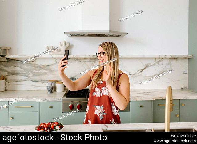 Female nutrient expert waving hand during video call through smart phone in kitchen