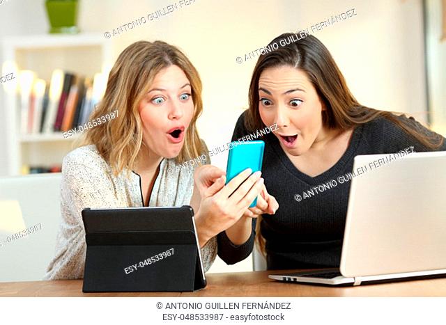 Front view portrait of two amazed friends using multiple devices at home