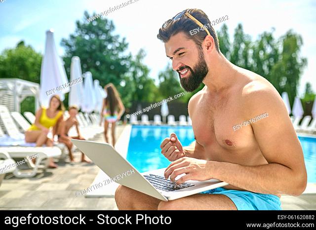 Focused excited bearded dark-haired young man sitting by the swimming pool and staring at his laptop