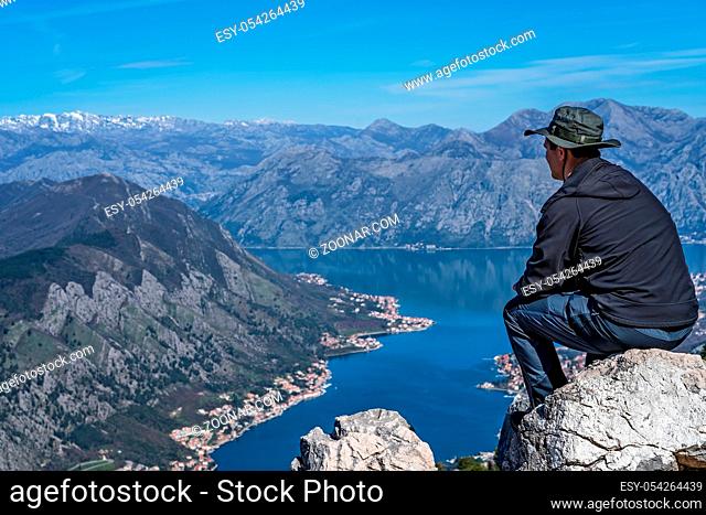 Tourist with a hat sitting on a large boulder and admiring the stunning landscape of the Bay of Kotor in Montenegro as seen from the road to Lovcen National...