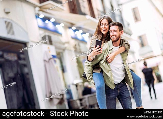 Cheerful woman taking selfie through smart phone while piggybacking on boyfriend