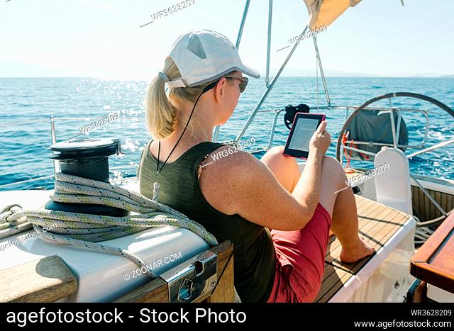 Woman sitting on deck of a yacht enjoying reading in her ebook. Travel and vacation concept