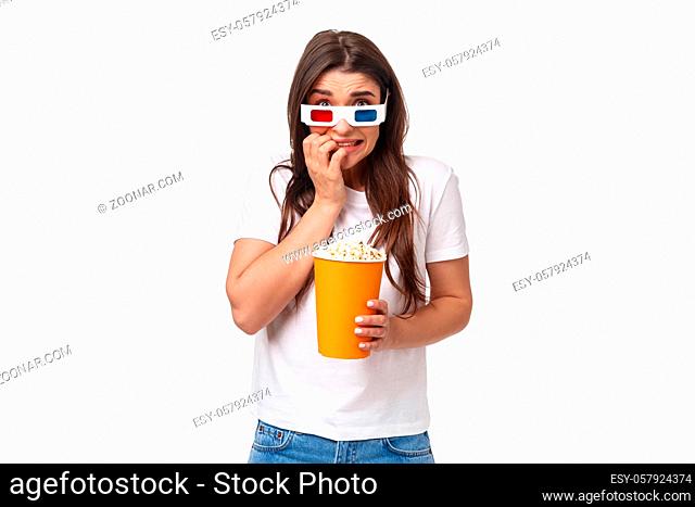 Entertainment, fun and holidays concept. Portrait of scared young cute girl in 3d glasses, eating popcorn, stooping and biting fingers as being scared of horror...